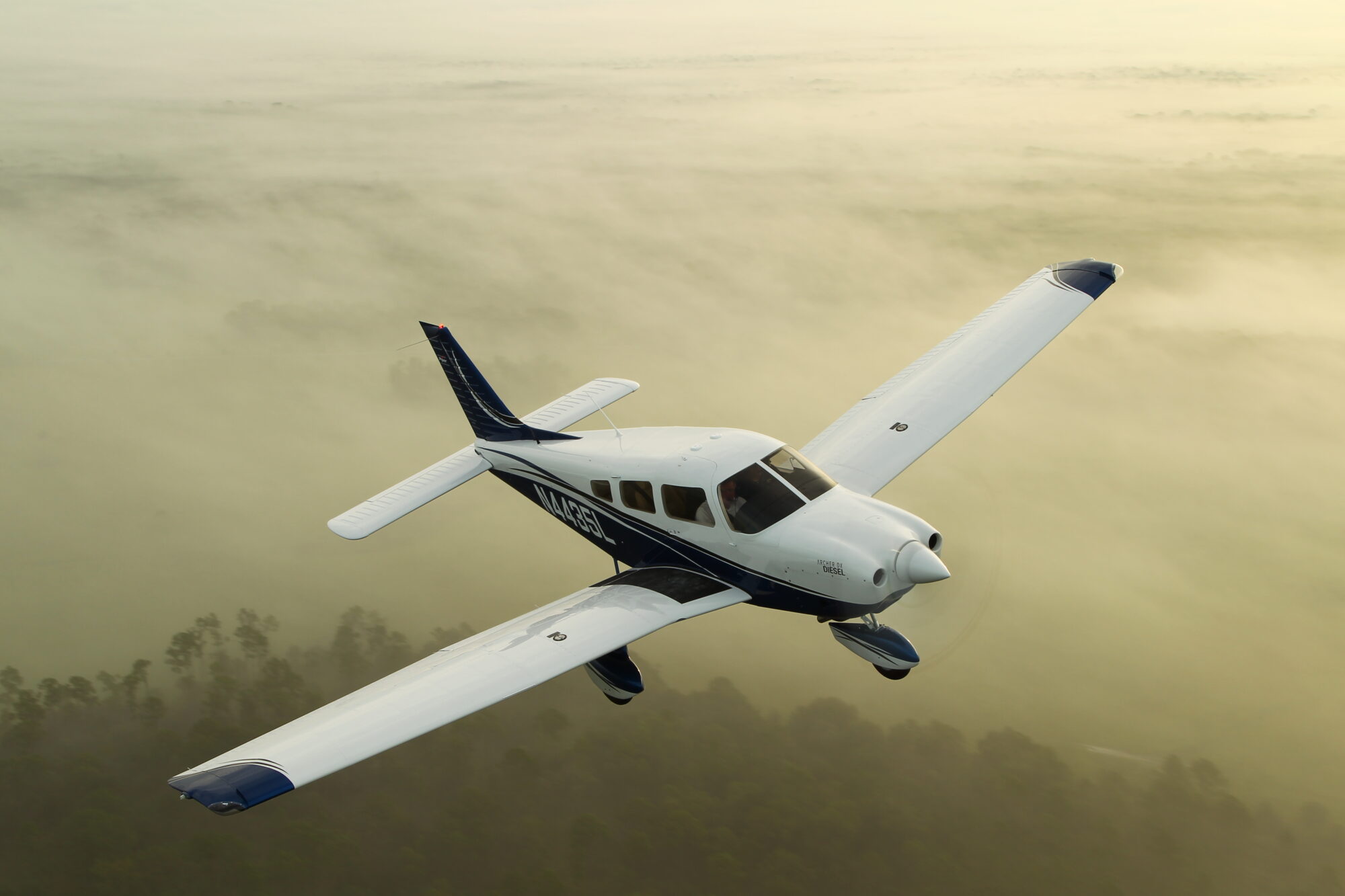 A Piper Archer DX airplane flying over a misty, forested landscape with sunlight illuminating the aircraft and the clouds below.