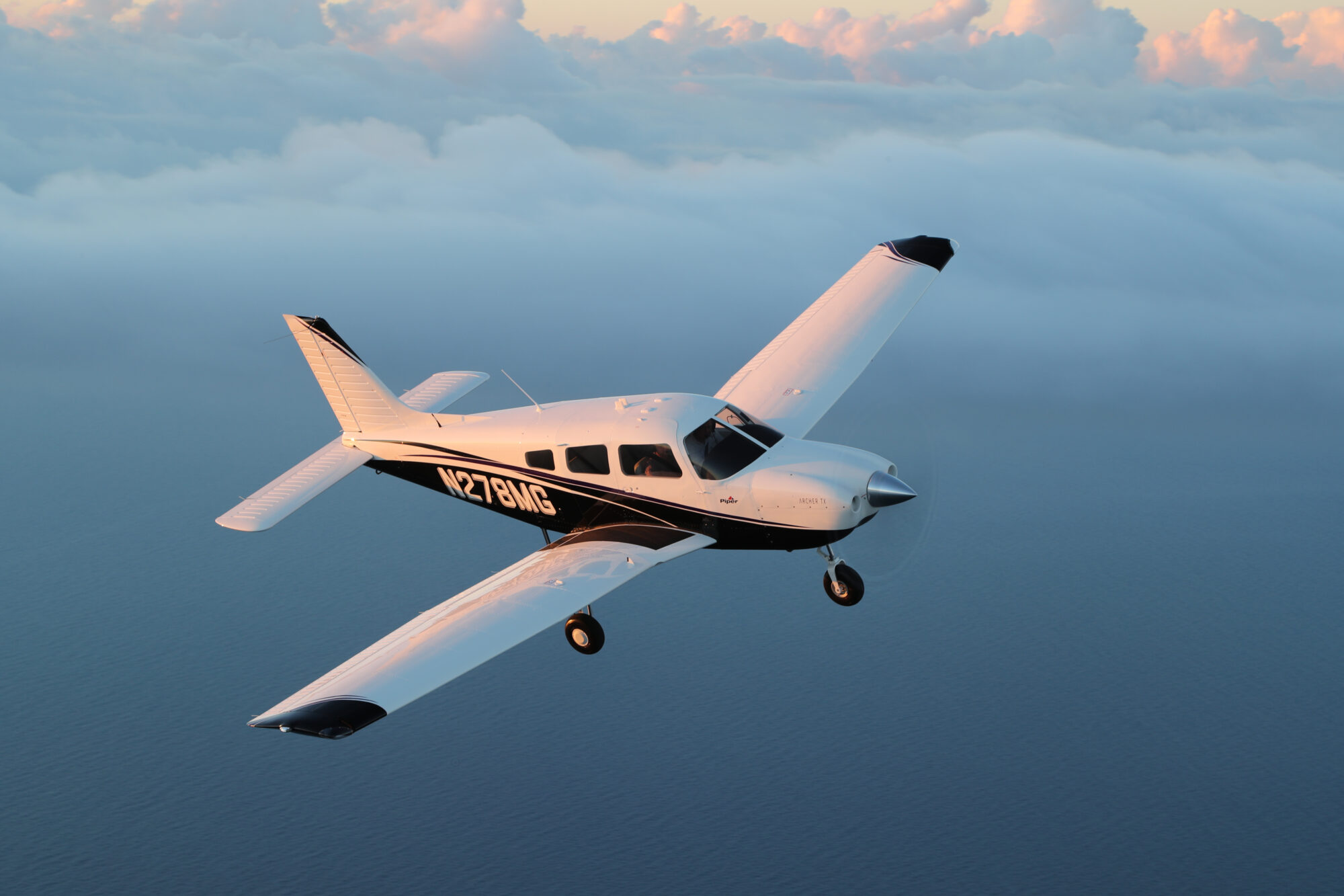 A Piper Archer TX airplane flying over a serene ocean landscape with a backdrop of soft, fluffy clouds lit by the sunrise.