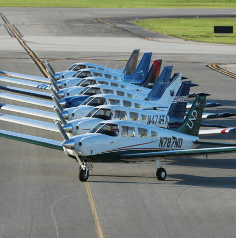 A fleet of Piper Archer aircraft lined up on an airport tarmac, showcasing various tail designs and school logos.