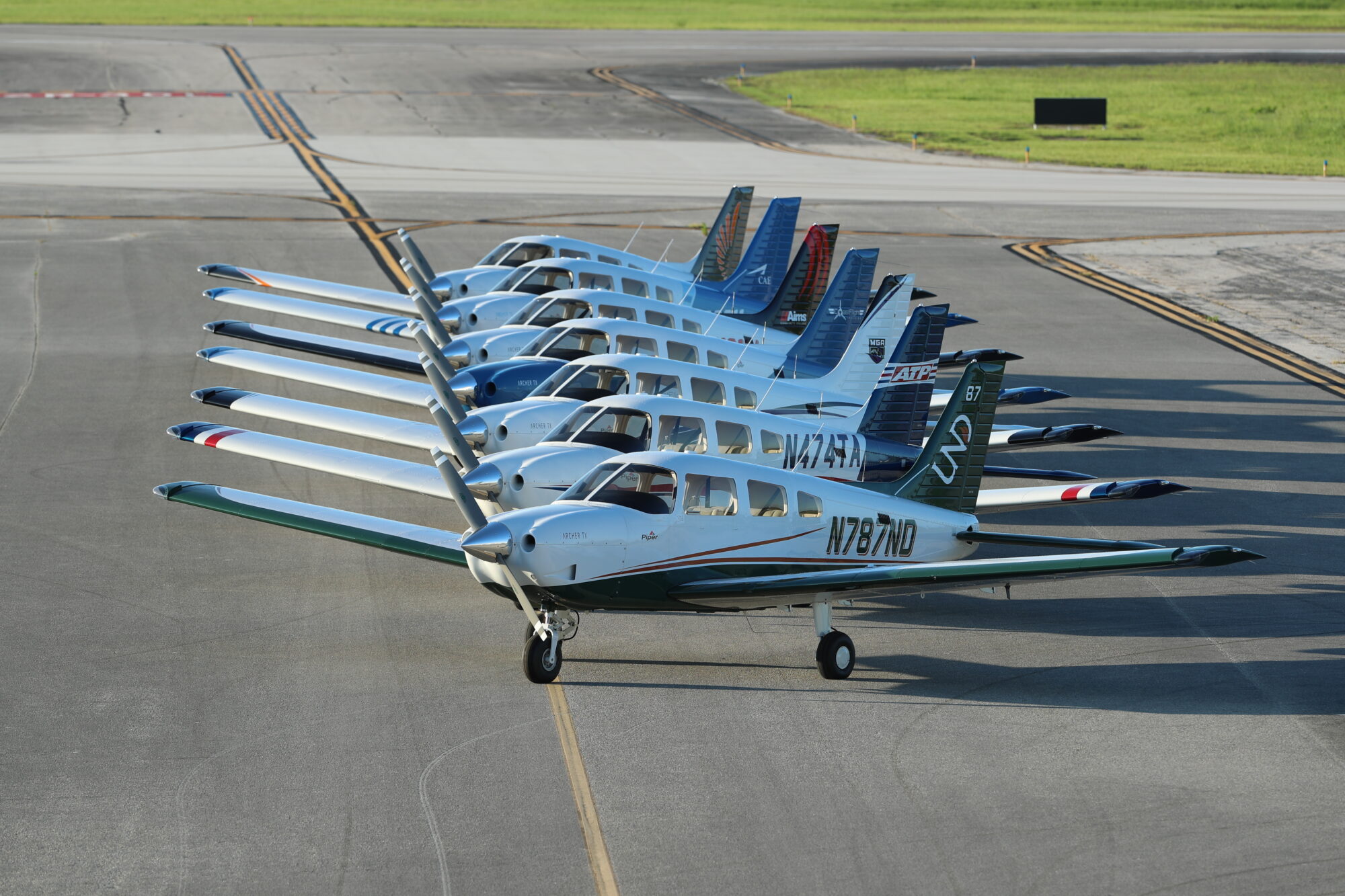 A fleet of Piper Archer aircraft lined up on an airport tarmac, showcasing various tail designs and school logos.