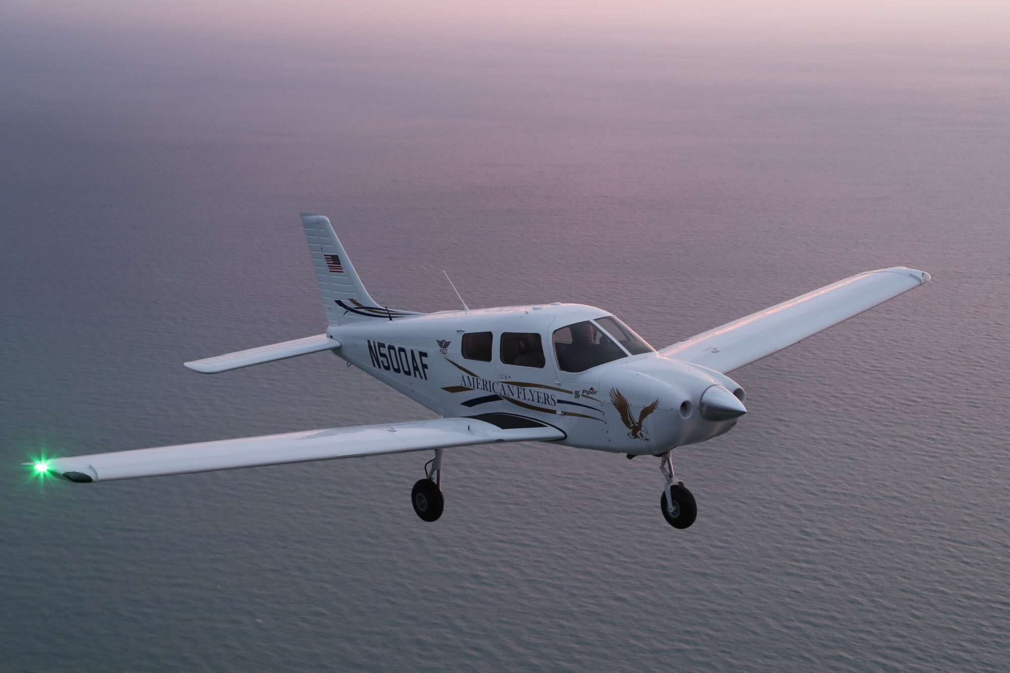 A Piper Pilot 100i airplane flying over an ocean during the evening with subtle pink hues in the sky.