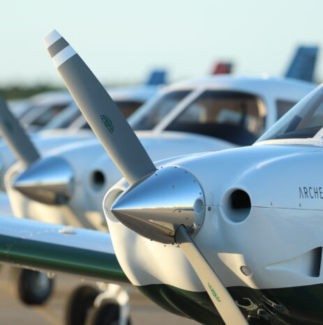 Close-up view of the propeller and nose of a Piper Archer TX with several other training aircraft lined up on the tarmac in the background.