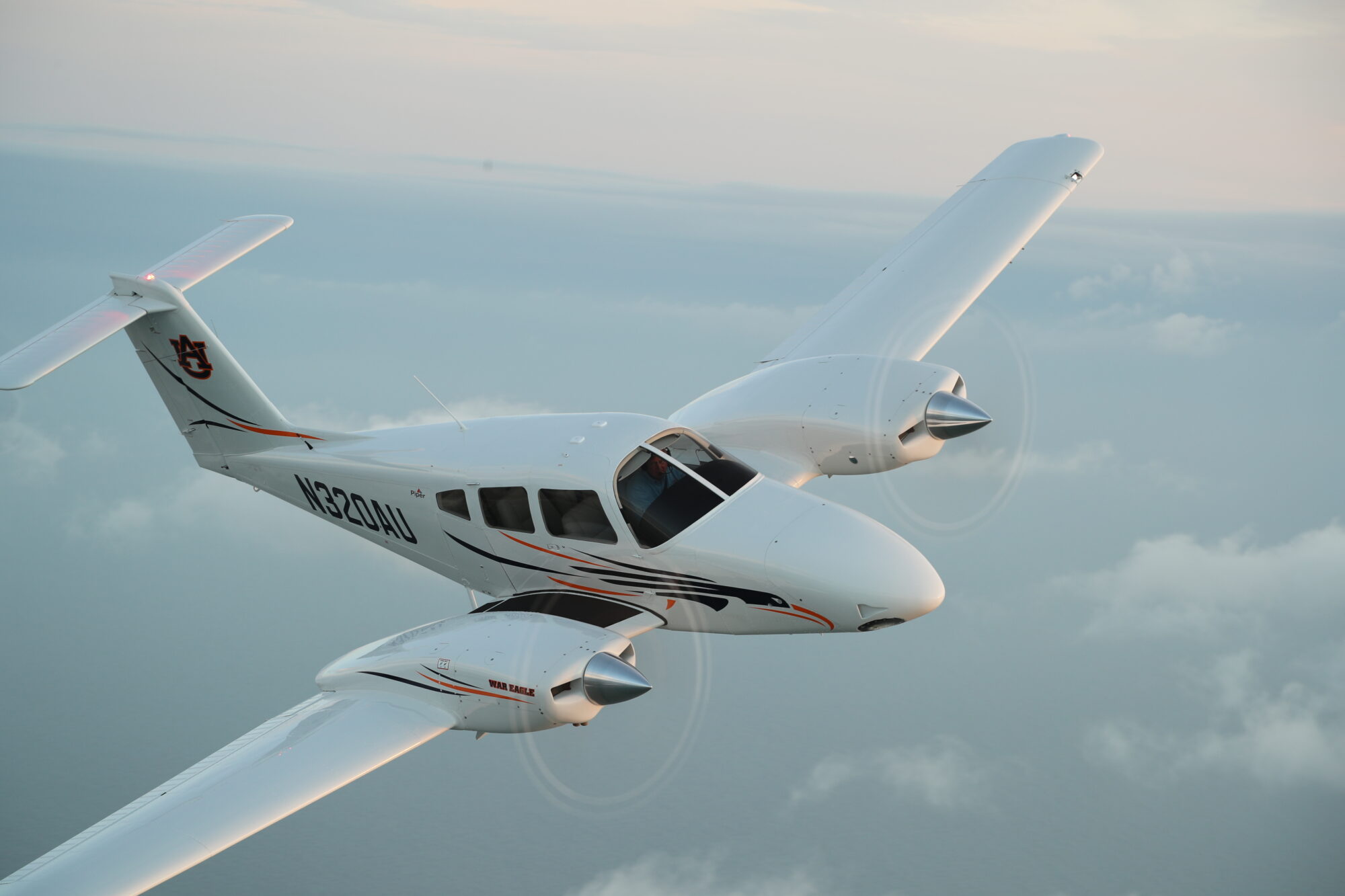 Piper Seminole multi-engine training aircraft flying over a coastal landscape with clouds below and the horizon in the background.